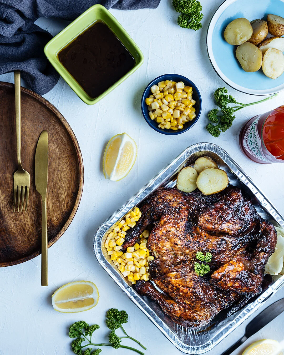 Roast chicken with potatoes, corn, and sauce surrounded by lemon wedges and parsley on a white table.