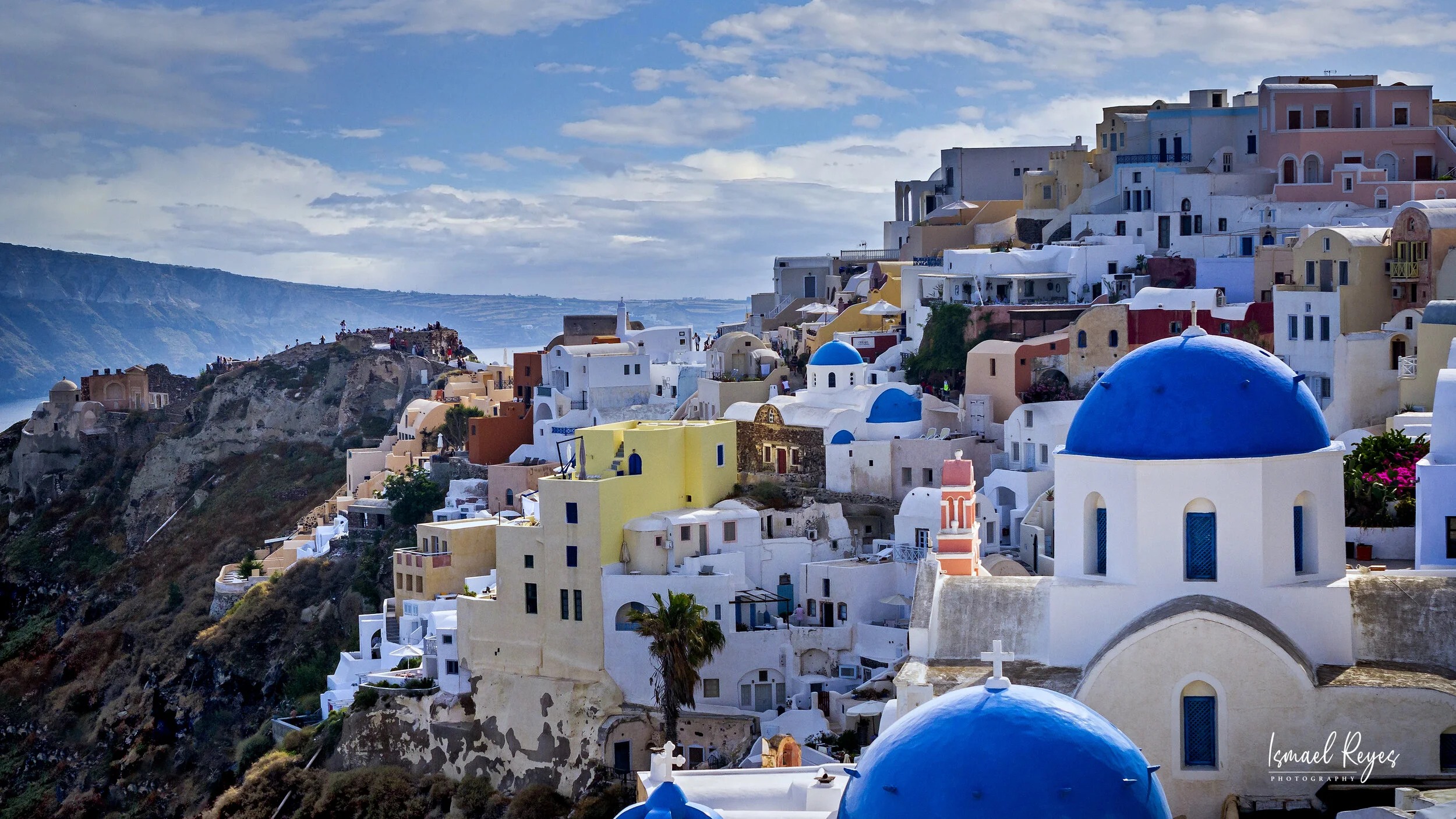 Colorful buildings with blue domes on a hillside in Santorini, Greece, under partly cloudy sky.