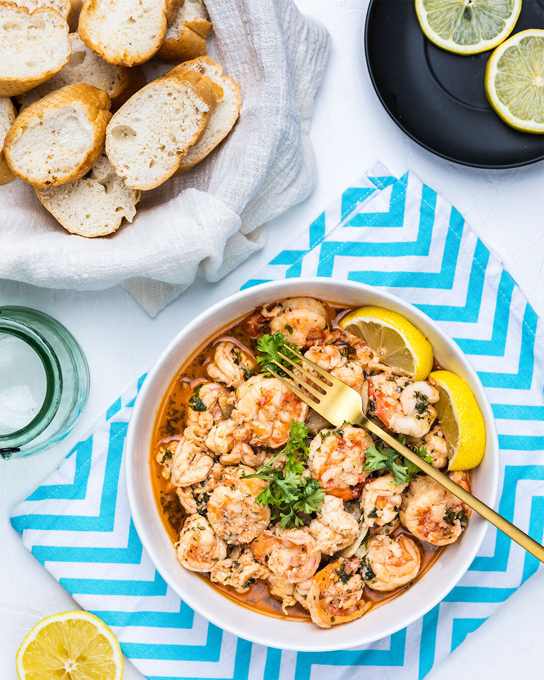 A bowl of cooked shrimp garnished with cilantro and lemon wedges, accompanied by slices of baguette and lemon slices on a black plate, set on a white surface with a blue and white patterned cloth nearby.