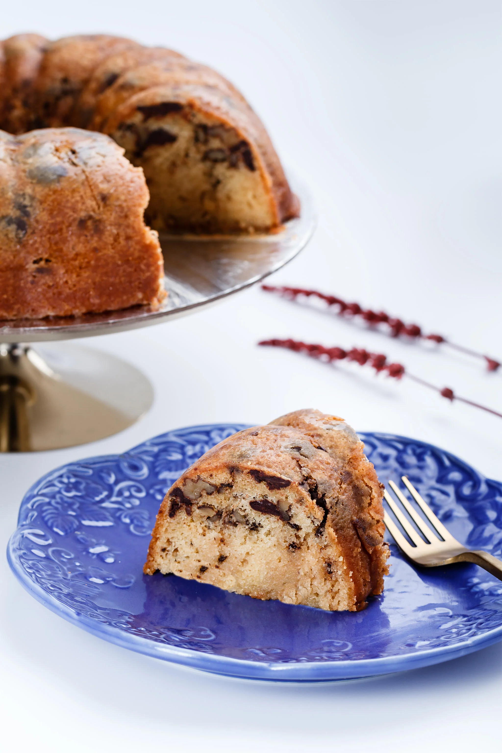 A slice of chocolate chip cookie cake on a blue plate with a fork, with the remaining cake on a silver cake stand in the background.