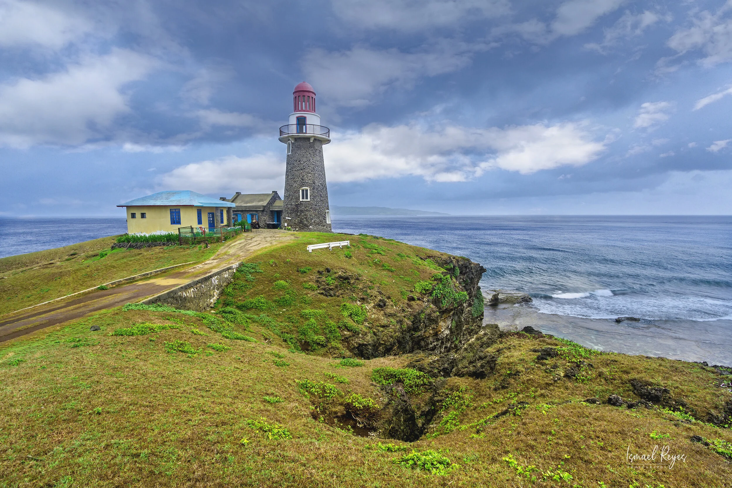A lighthouse on a grassy cliff overlooking the ocean under a partly cloudy sky.