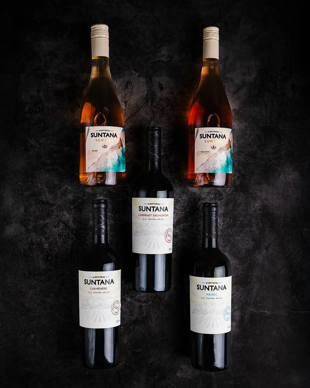 Five bottles of wine on a dark textured surface, with two bottles of Suntana Summer rosé wine at the top, and three bottles of Suntana red wines (Carménère, Cabernet Sauvignon, and Malbec) at the bottom.