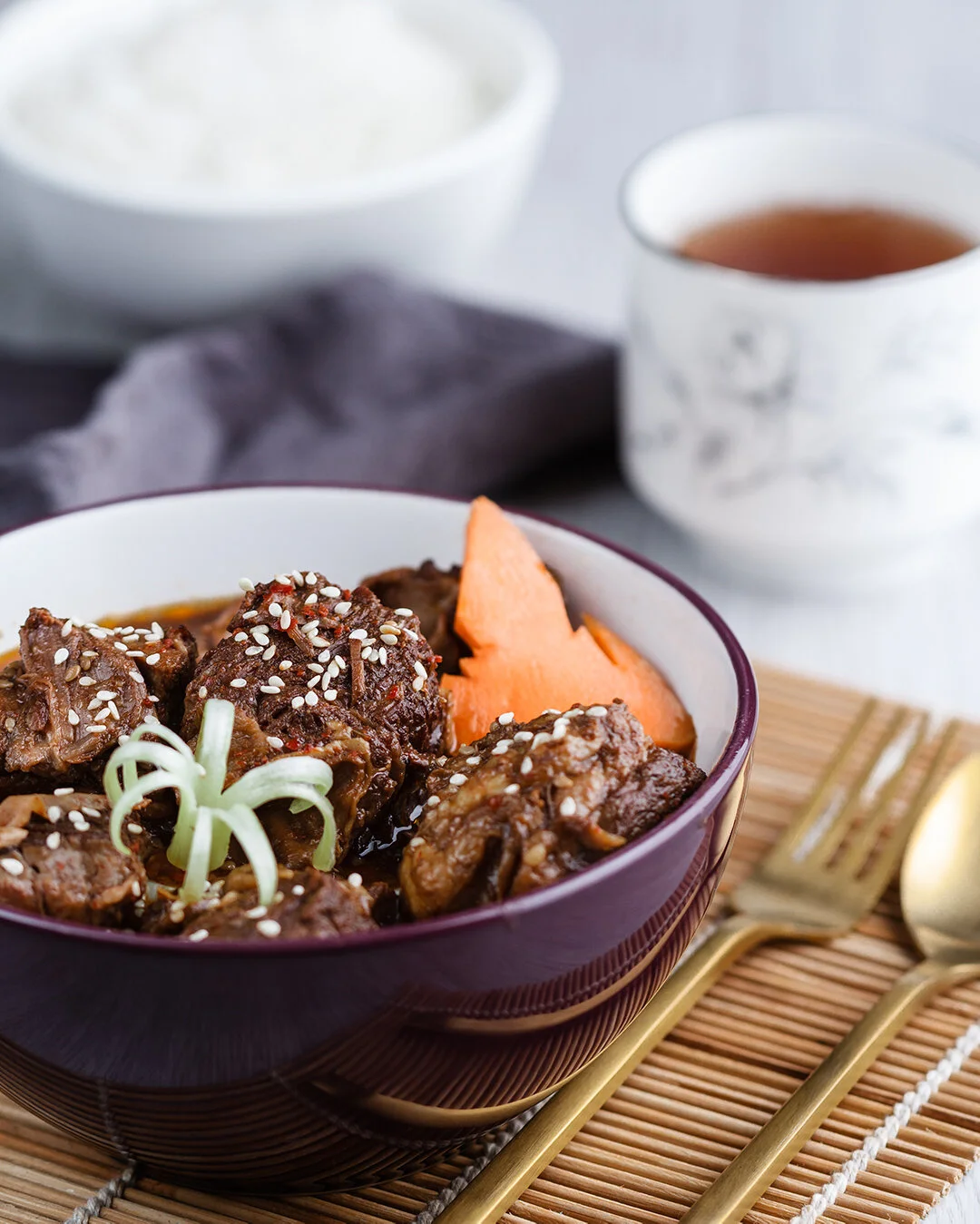 Bowl of beef stew garnished with sesame seeds and green onion, with a slice of carrot, on a bamboo mat with a fork and spoon nearby. In the background, a cup of tea and a bowl of rice are visible.