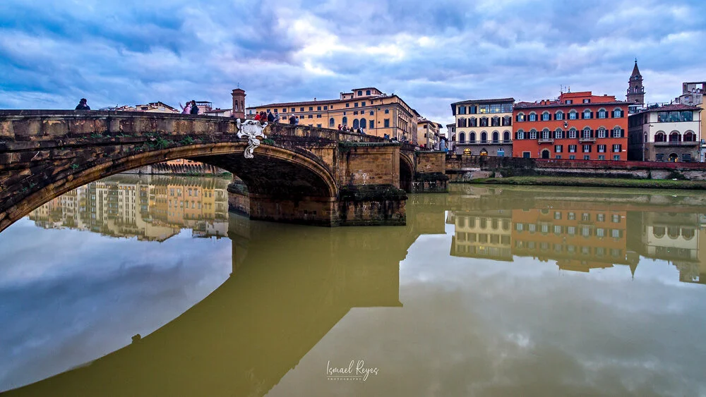 A river with a stone bridge, buildings reflected in the water, and cloudy sky overhead.