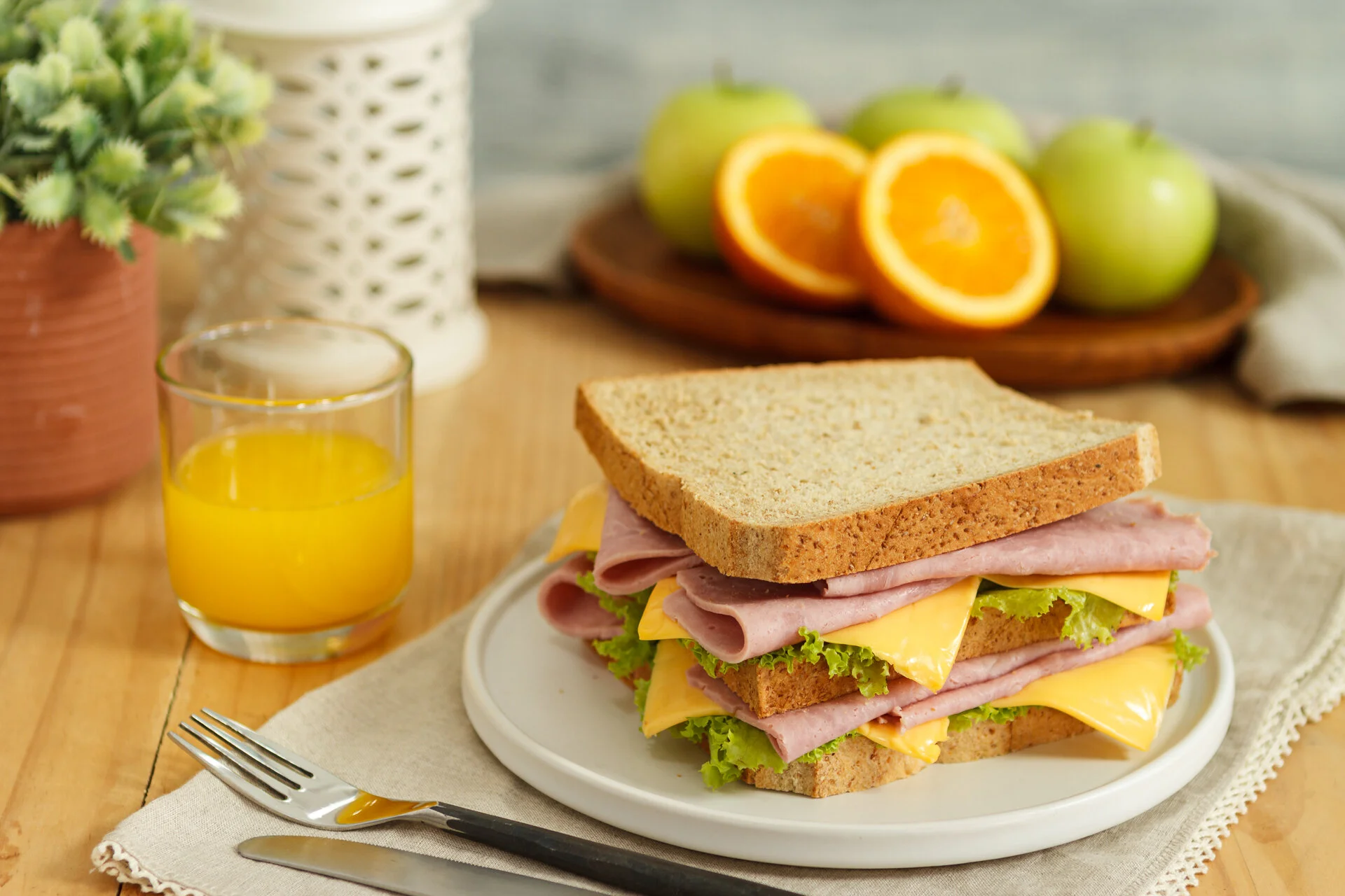 A plate with a sandwich made of whole wheat bread, slices of ham, cheddar cheese, and lettuce. A glass of orange juice is beside the plate. In the background, there are green apples and orange halves on a wooden tray, and a potted plant with green fo