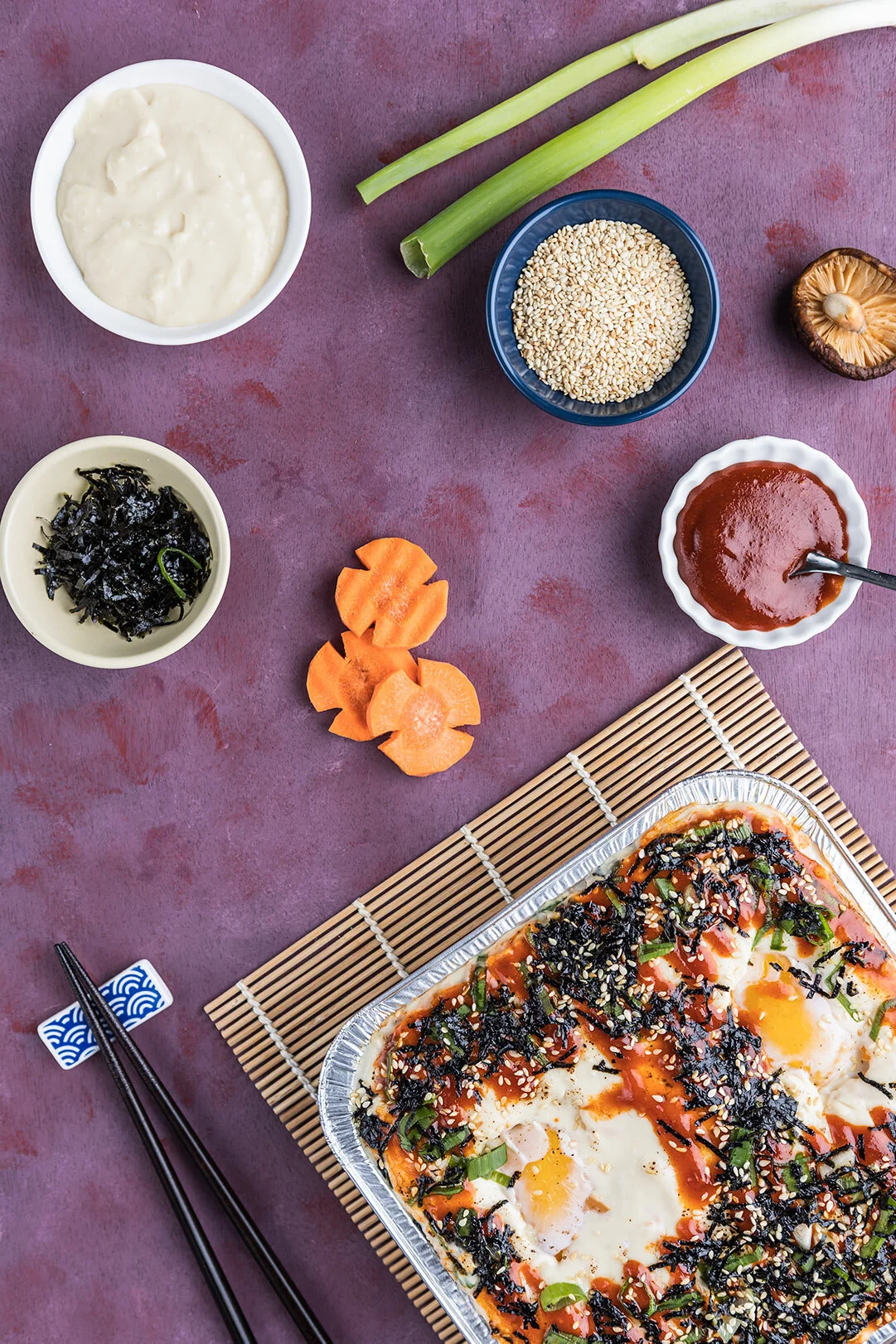 Korean dish with a baked egg and rice topped with seaweed, sauce, and sesame seeds, surrounded by bowls of sauce, seaweed, sesame seeds, green onion, bamboo steamer with mushrooms, and sliced carrots, with chopsticks nearby.