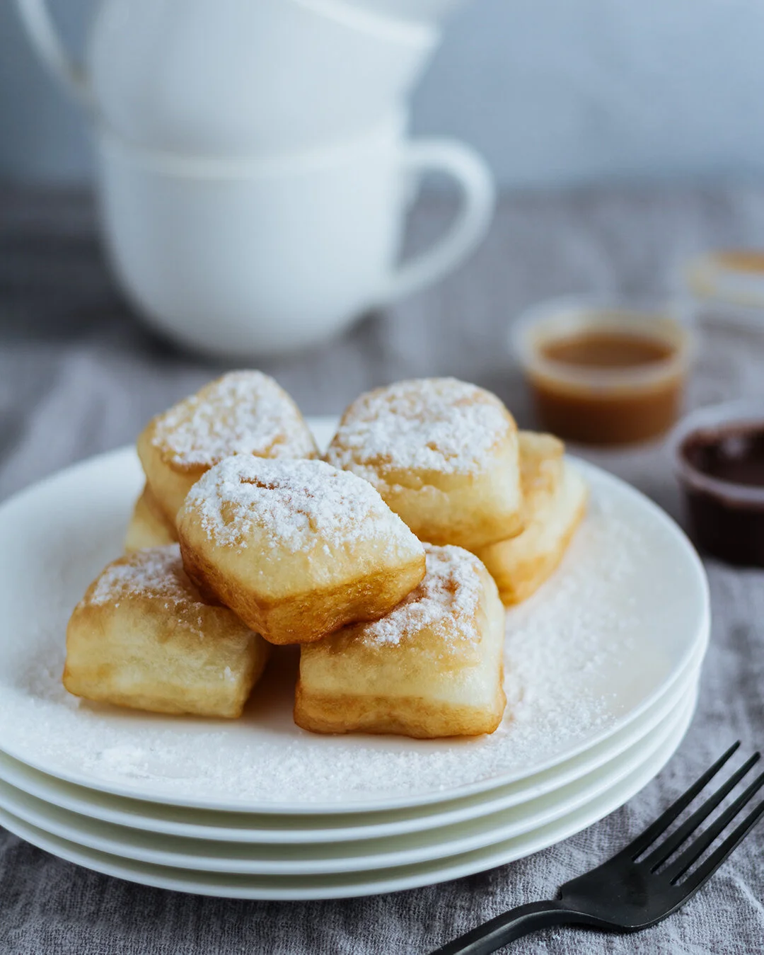 Stack of fried dough squares dusted with powdered sugar on a white plate, with syrup and a coffee mug in the background.