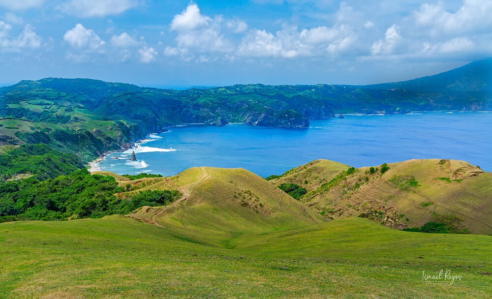Lush green rolling hills leading to a blue ocean with cliffs in the background under a partly cloudy sky.