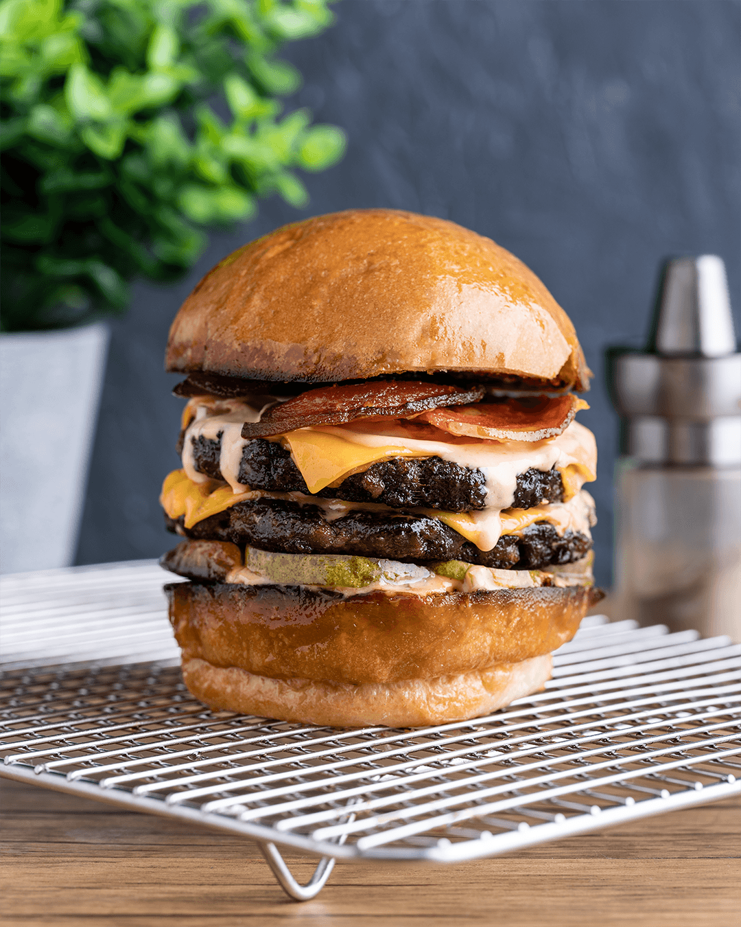 A large, multi-layered cheeseburger with pickles, lettuce, melted cheese, beef patties, bacon, and a toasted bun, placed on a wire cooling rack.