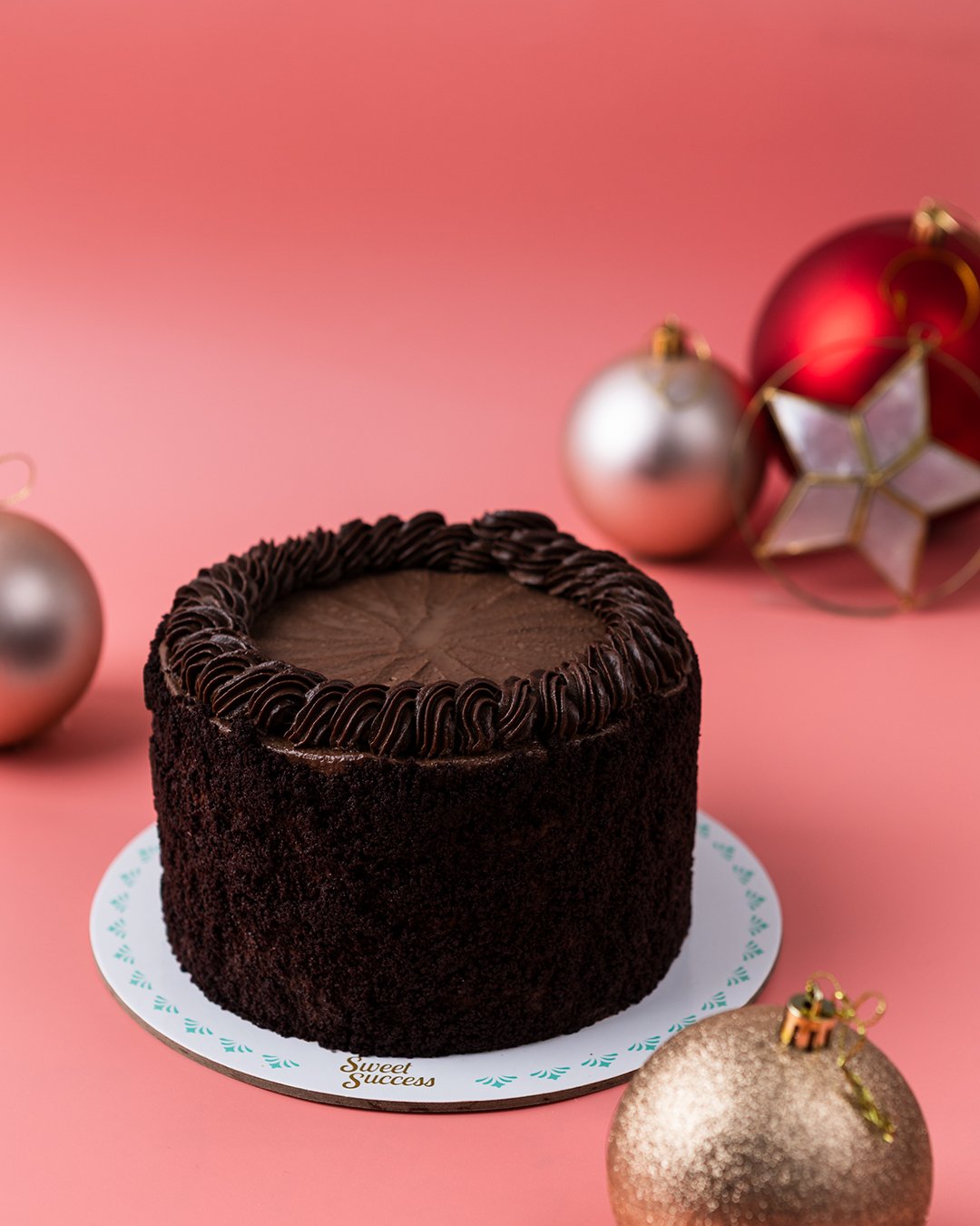 A chocolate cake with decorative frosting on top, placed on a white plate, surrounded by Christmas ornaments on a pink background.