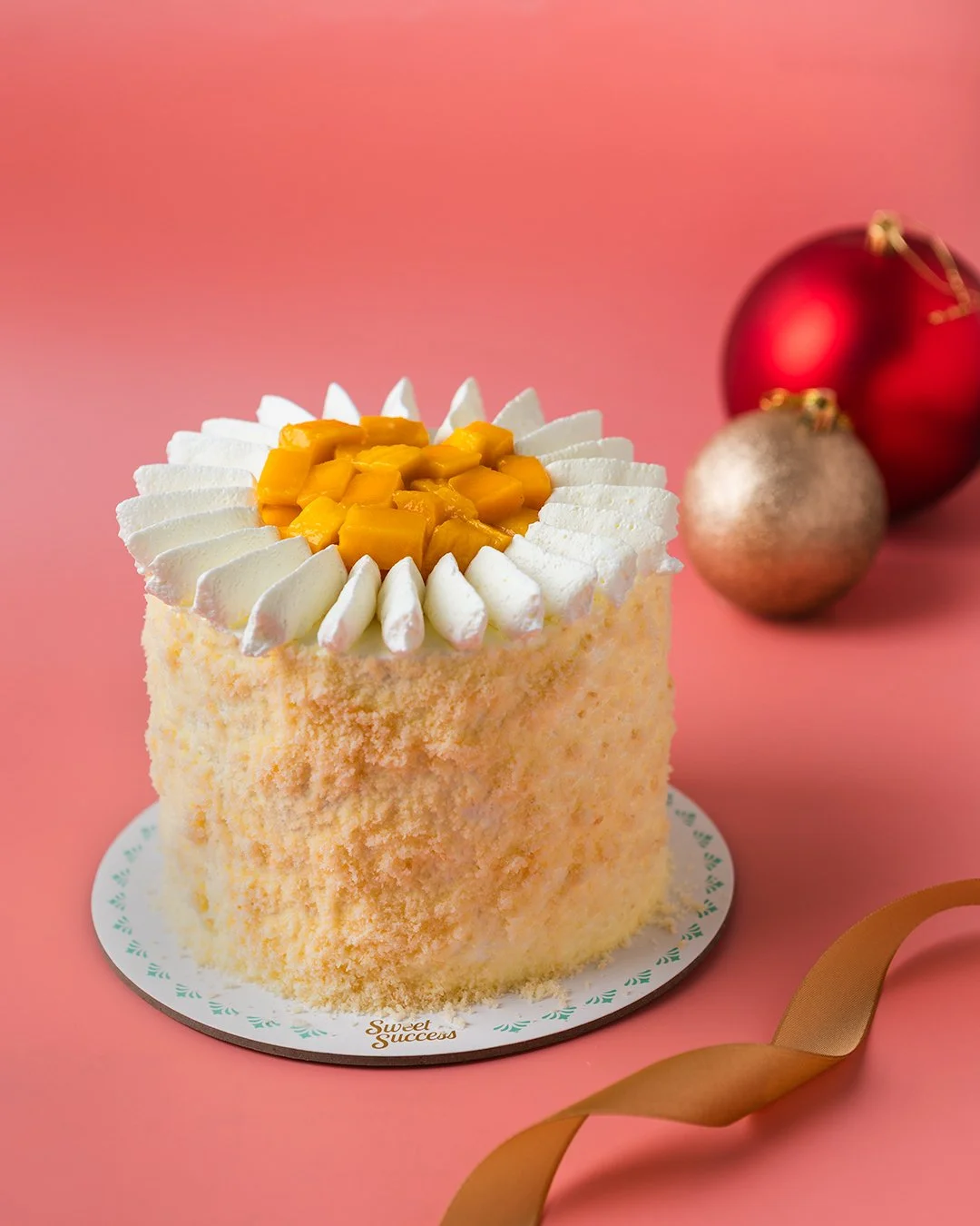 A small round cake with white frosting, topped with canned mango pieces and white frosting decoration. The cake sits on a white cake board with "Sweet Success" written on it, set against a pink background with red and gold Christmas ornaments and a g