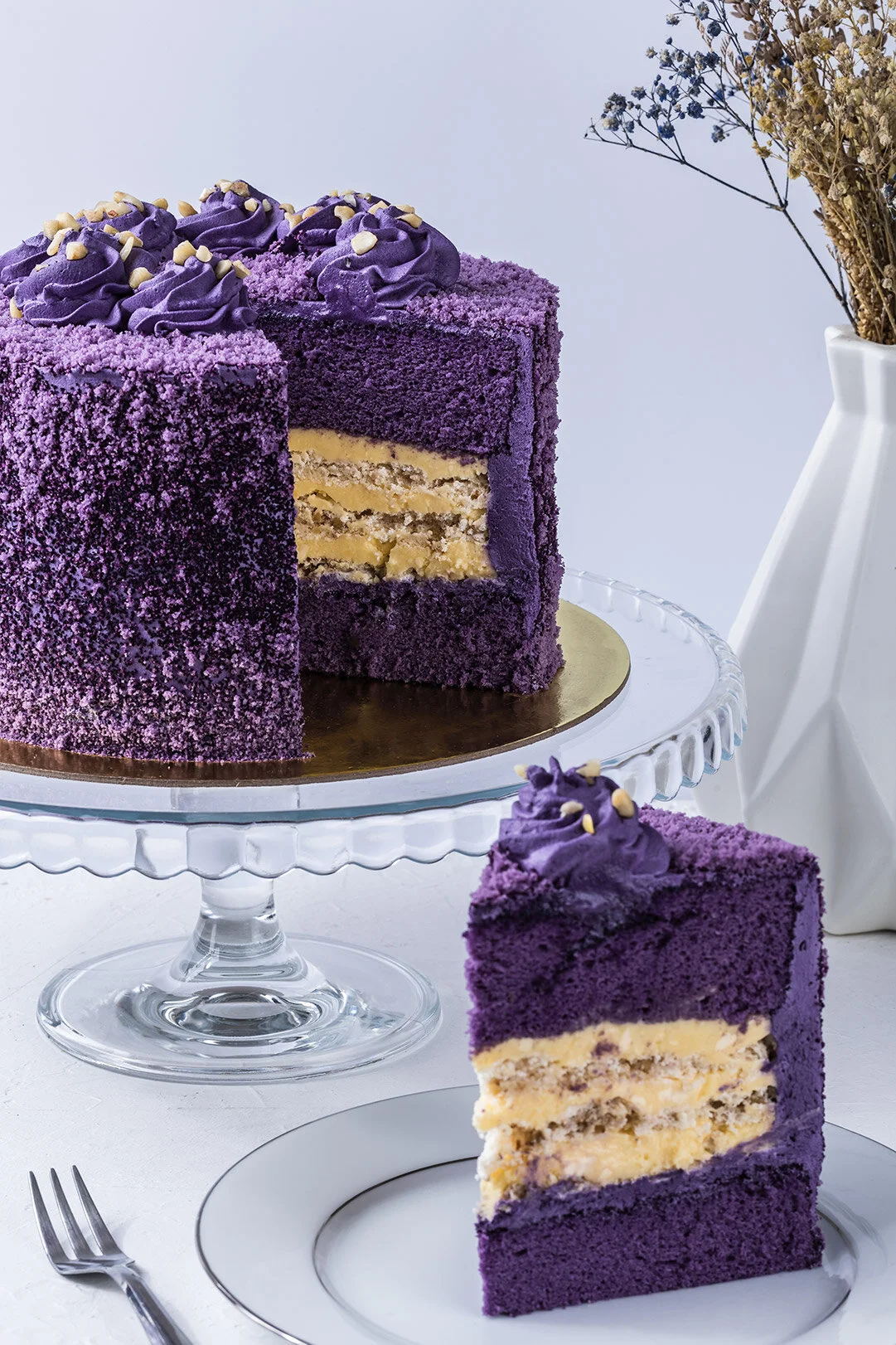 A purple layered cake with purple frosting and sprinkles on top, displayed on a glass cake stand. A slice of the cake is cut and placed on a white plate nearby, showing the cake's layers and cream filling.