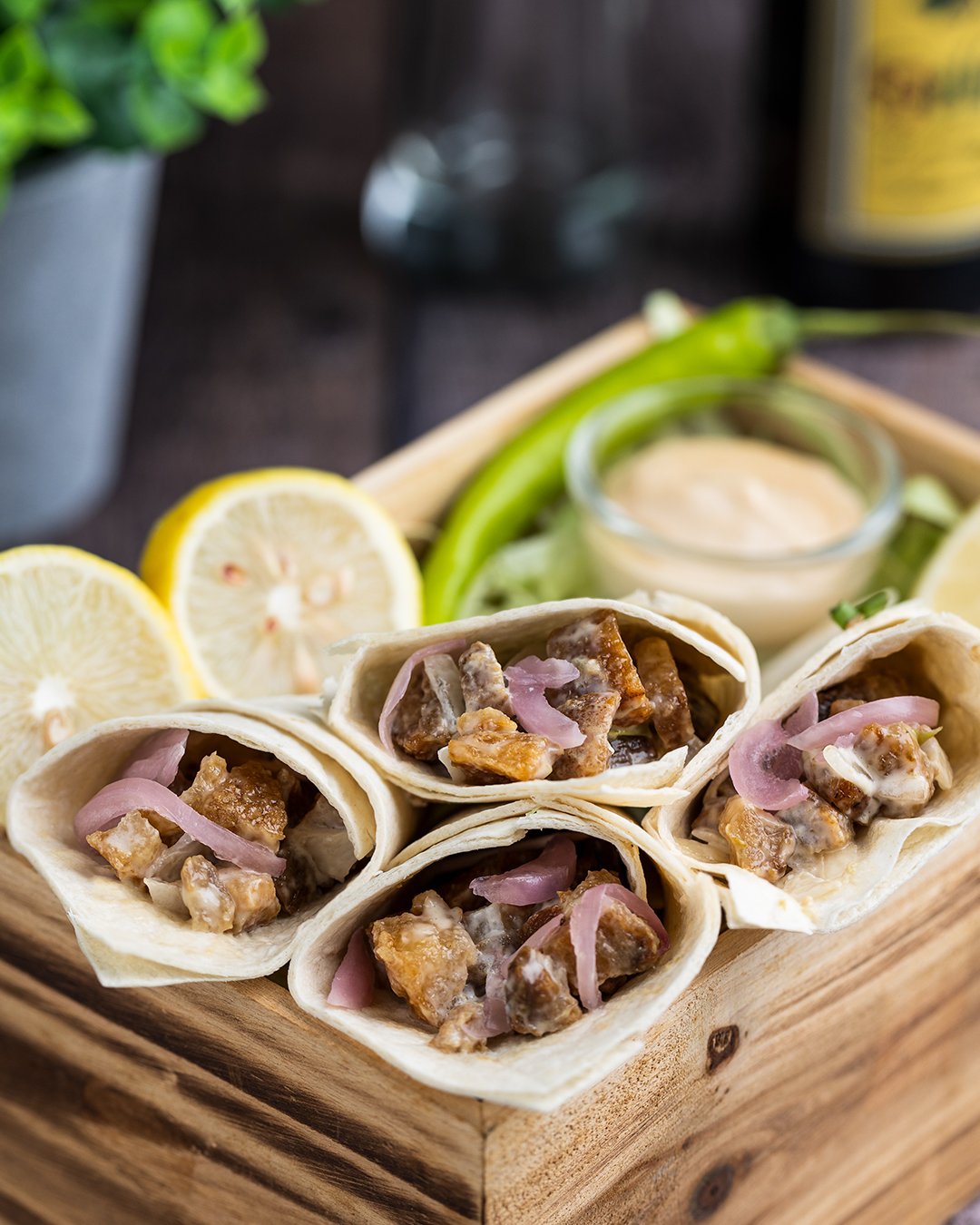 Close-up of five mini tacos filled with pork, onions, and sauce on a wooden tray. In the background, there are lemon slices, cucumber, a small bowl of dipping sauce, and a jalapeño pepper, with a blurred glass and a yellow bottle.