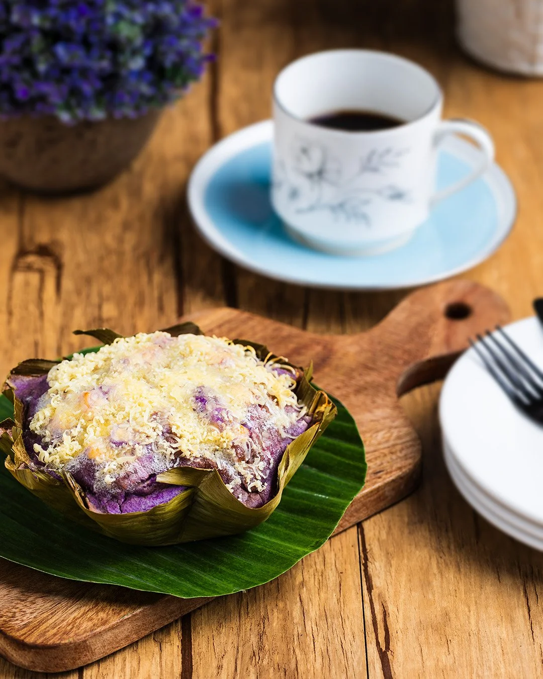 A purple sweet potato dish topped with grated cheese on a banana leaf, on a wooden serving board, with a cup of black coffee in a white cup with a floral design, a white plate with a fork, and a blue flower in the background.