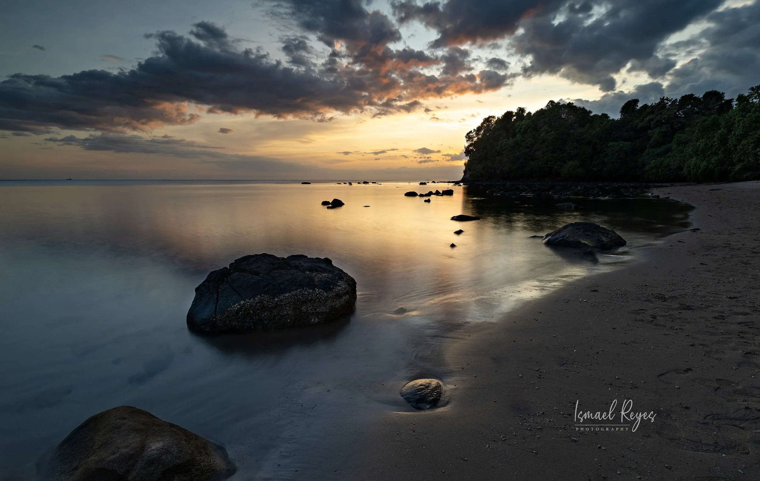 A tranquil beach scene at sunset with rocks in the shallow water, a sandy shoreline, and a wooded hill in the distance under a partly cloudy sky.