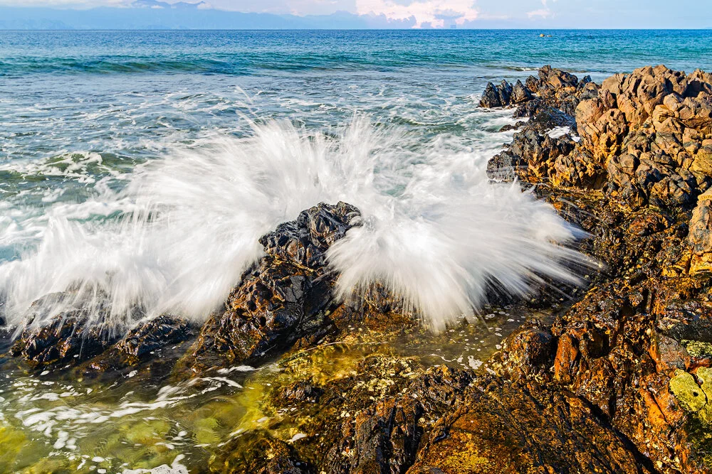 Waves crashing against rocks on a seaside shore with a clear blue sky in the background.