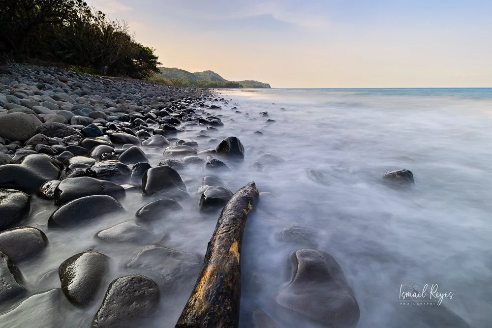 A rocky shoreline with smooth black stones and a large driftwood log, with calm ocean waves washing over the rocks, and a distant tree-lined headland under a partly cloudy sky.