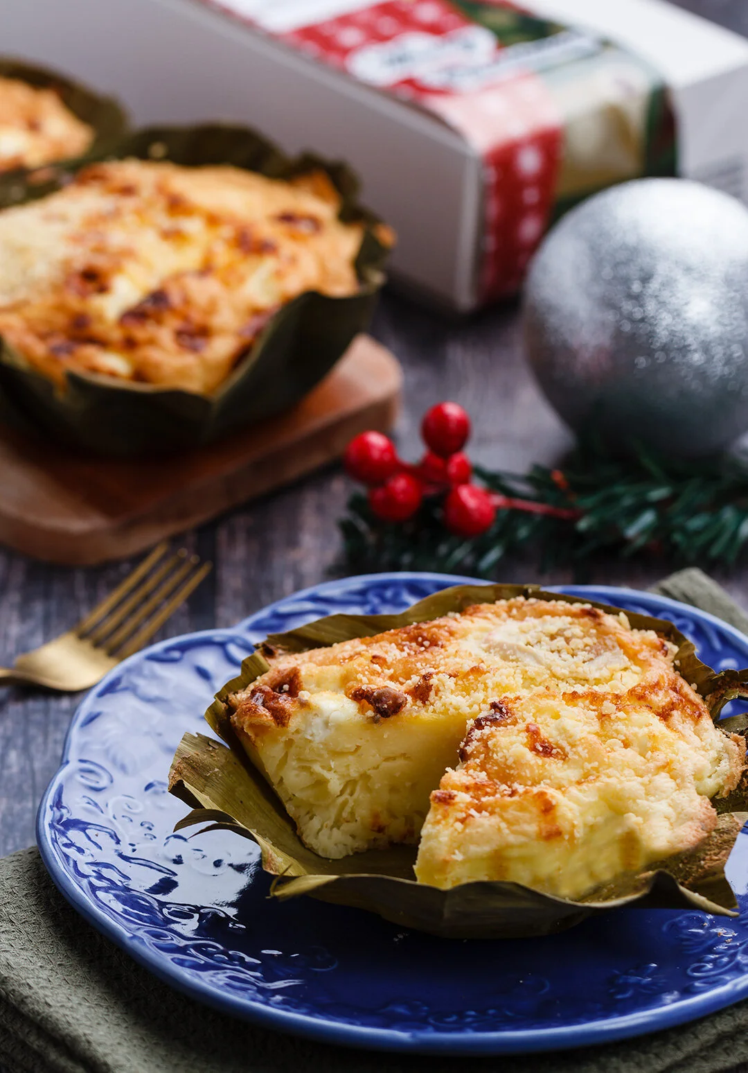 A slice of creamy potato casserole topped with cheese on a blue plate, with a gold fork beside it, and a whole casserole dish in the background, decorated for Christmas with red berries, green foliage, a silver ornament, and a Christmas box.
