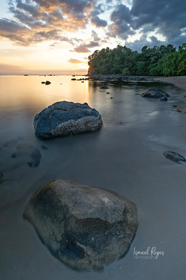 Sunset over a rocky beach with large stones in the foreground, calm water, and a lush green forest on the right side.