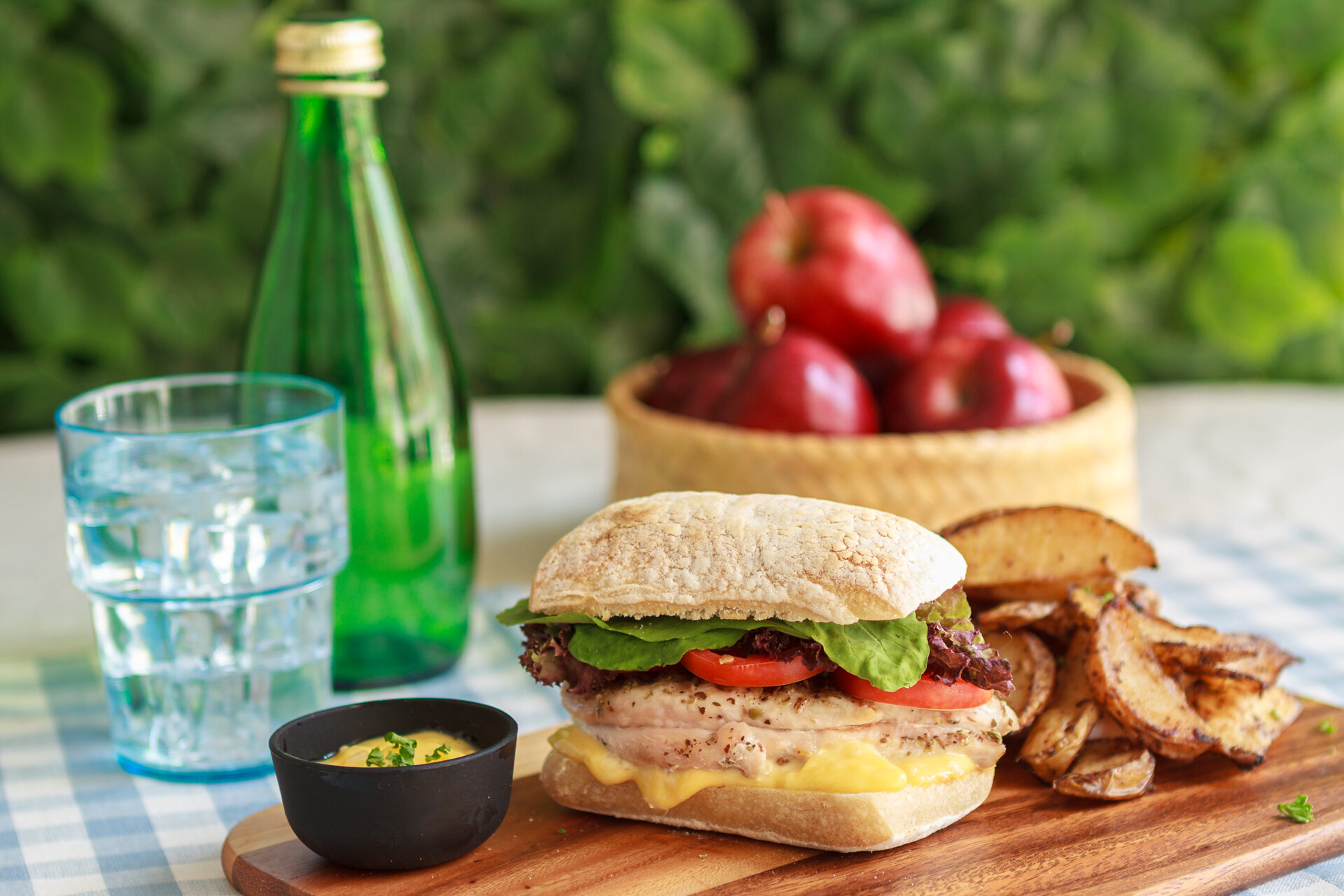A sandwich with lettuce, tomato, cheese, and meat on a bun, served with potato chips, a black dipping bowl, a bottle of water, and a glass of water on a picnic table with a green leafy background