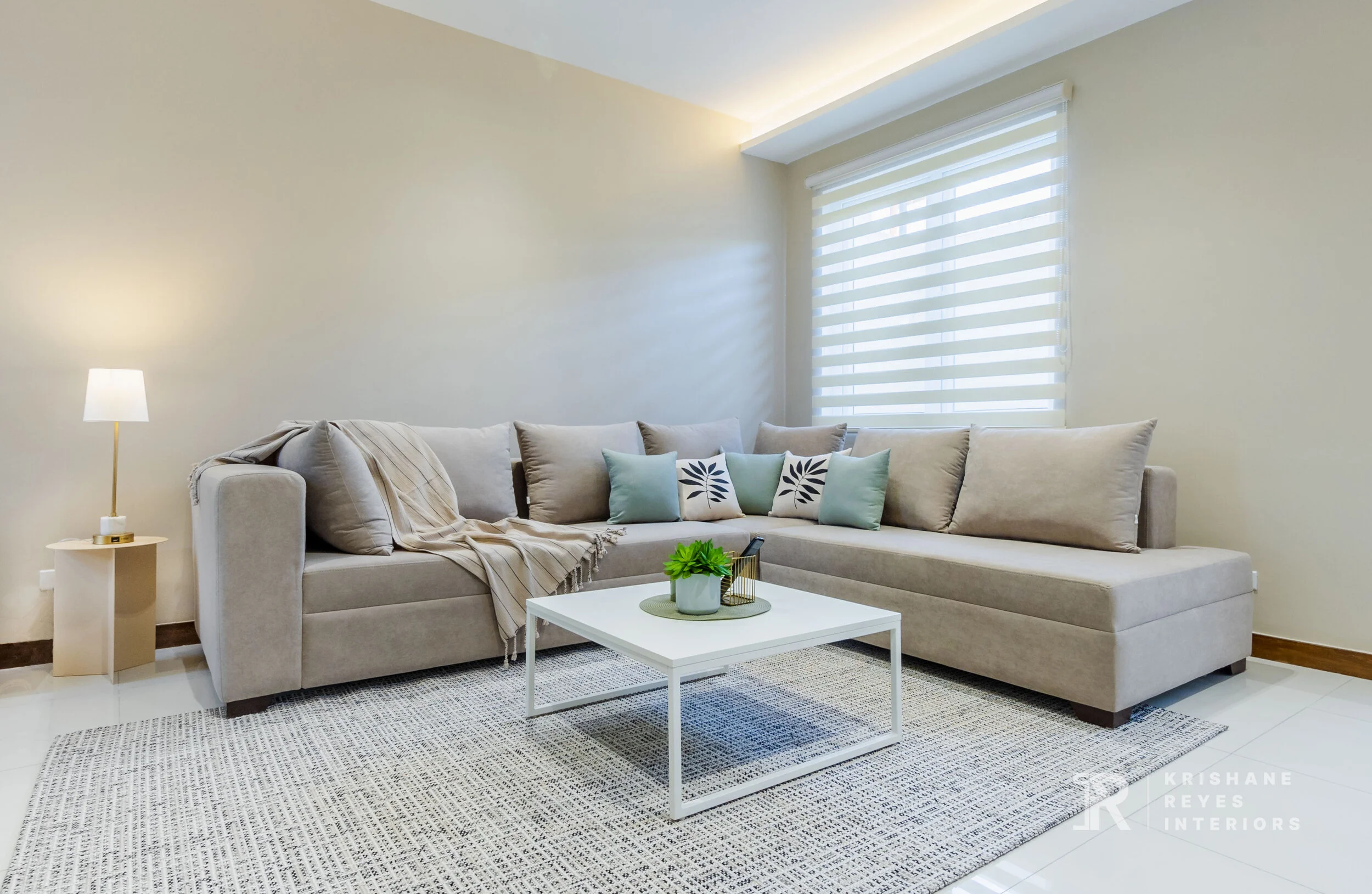 Bright living room with a light beige sectional sofa, decorative cushions, a small white coffee table with a green plant, and a side table with a lamp near a large window with horizontal blinds.
