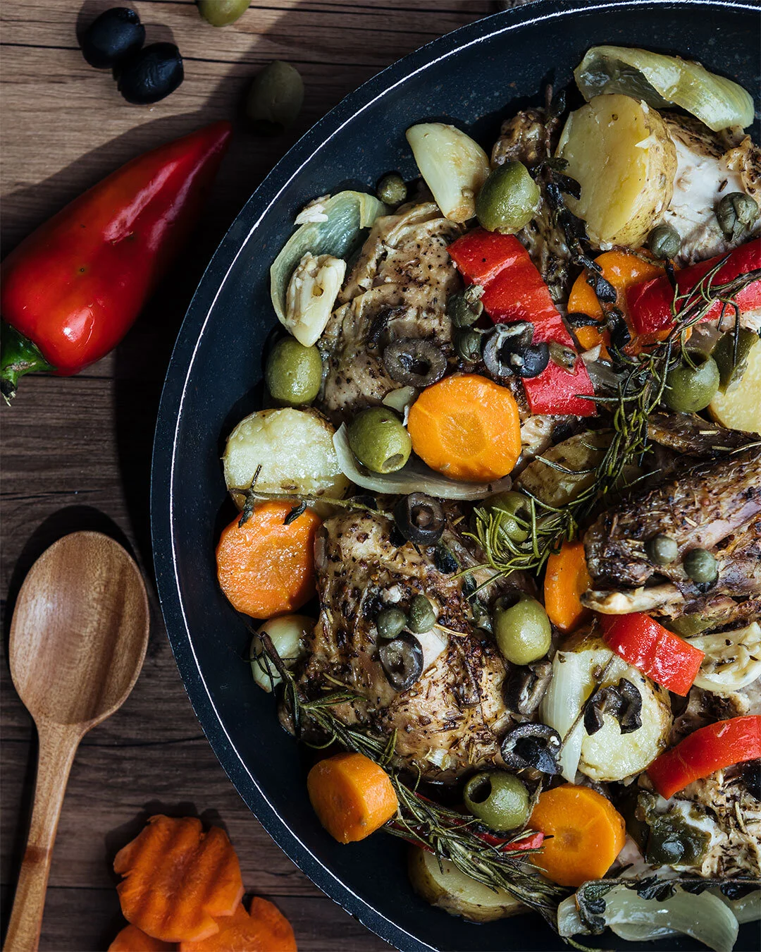 A roasted chicken with vegetables including potatoes, carrots, bell peppers, green olives, capers, and herbs in a black skillet. A red chili pepper and a wooden spoon are on the wooden surface beside the skillet.