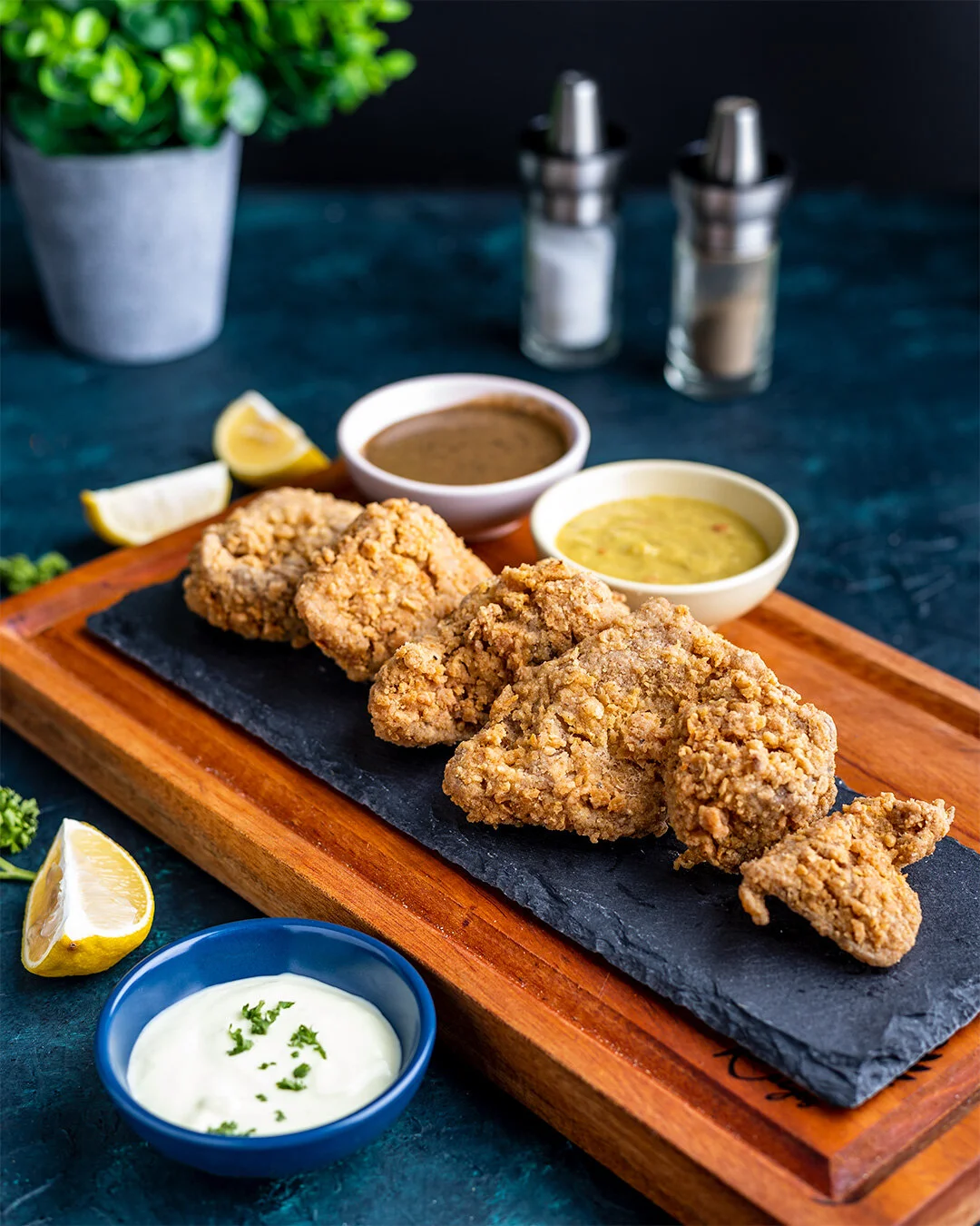 Plate of fried chicken drumsticks with lemon wedges, served with three bowls of dipping sauces on a wooden platter, with salt and pepper shakers, a potted plant, and lemon slices in the background.