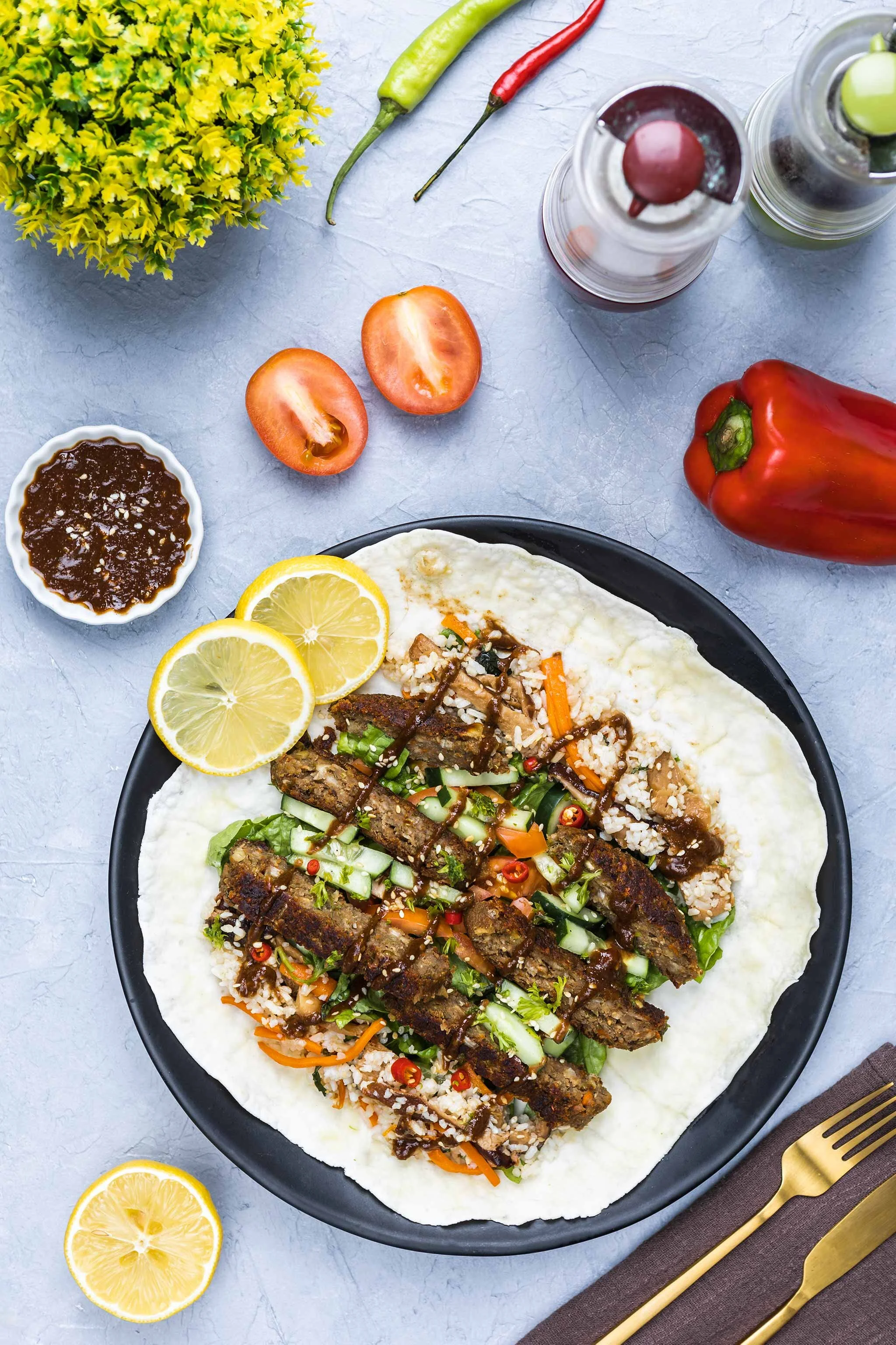 A plate of Asian-style lettuce wraps with grilled meat, chopped vegetables, and rice, garnished with lemon slices, with tomatoes, chili peppers, and soy sauce nearby, on a gray surface.