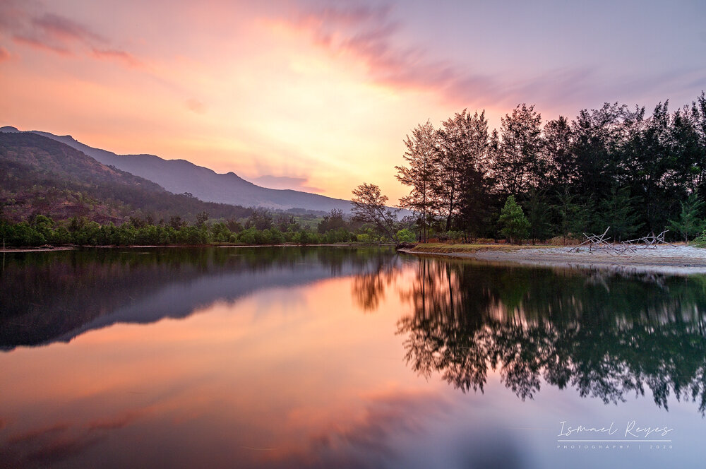 Sunset over a calm river with trees along the shoreline and mountains in the background, reflecting in the water.