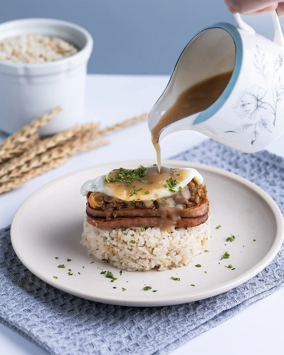 A plate with rice topped with a fried egg, ham, and sautéed mushrooms, with gravy being poured over it.