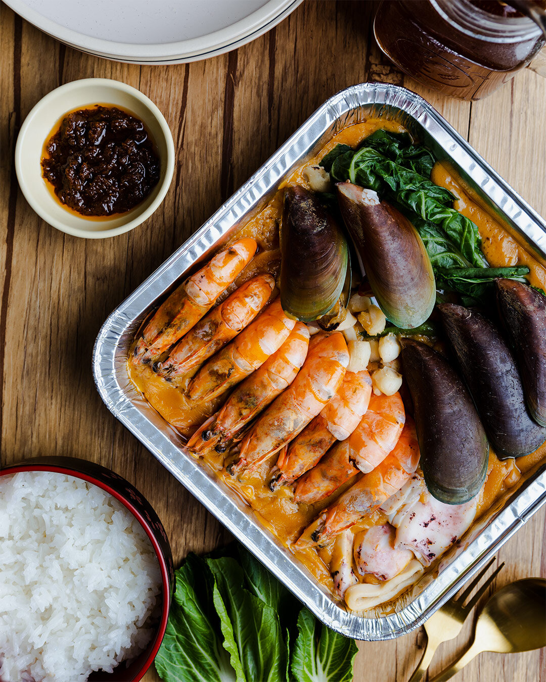 A seafood platter with prawns, mussels, squid, and vegetables in a foil container, served with a side of white rice, a small dish of dipping sauce, and a beverage on a wooden table.