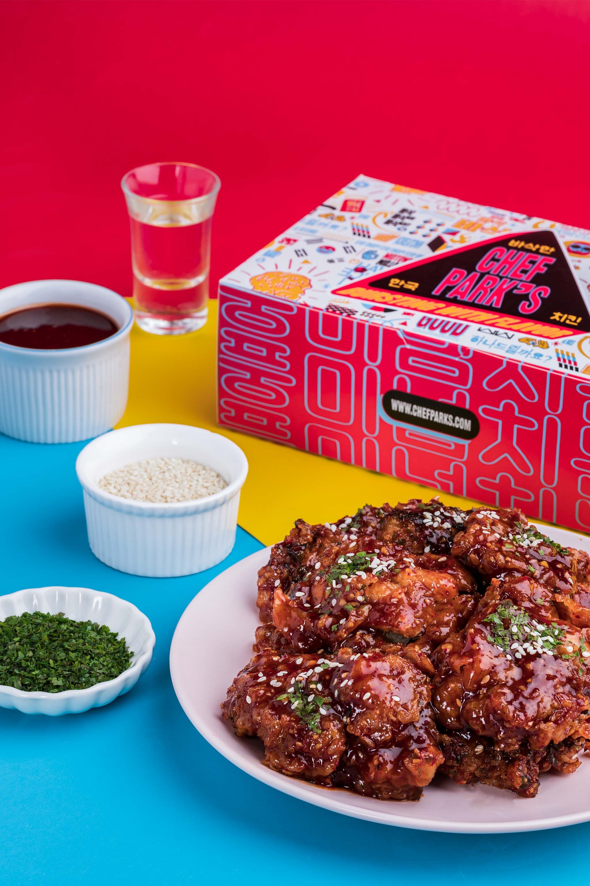 A white plate with glazed fried chicken pieces garnished with sesame seeds and chopped green onions, surrounded by small white bowls of herbs, sesame seeds, chopped green herbs, a glass of water, a cup of black coffee, and a box of Chef Park's Korean