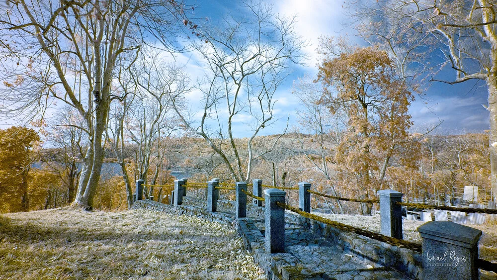 A wooden fence lining a stone pathway in a fall landscape with leafless trees, some with orange leaves, under a partly cloudy sky.