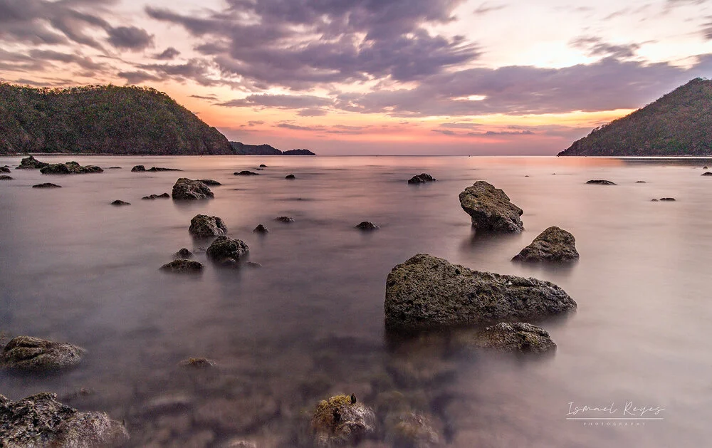 Coastal scene during sunset with rocks in calm water and hills on both sides.