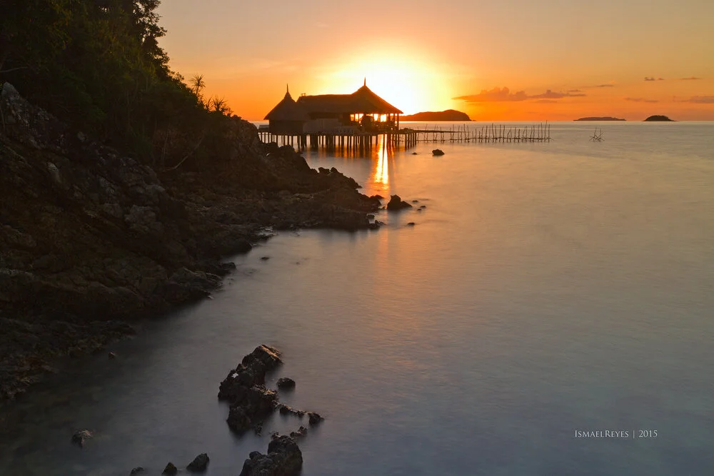 Sunset over a tropical seaside with clouds in the sky, a traditional wooden pier, and a hut over the water, with rocky shoreline and calm water reflections.