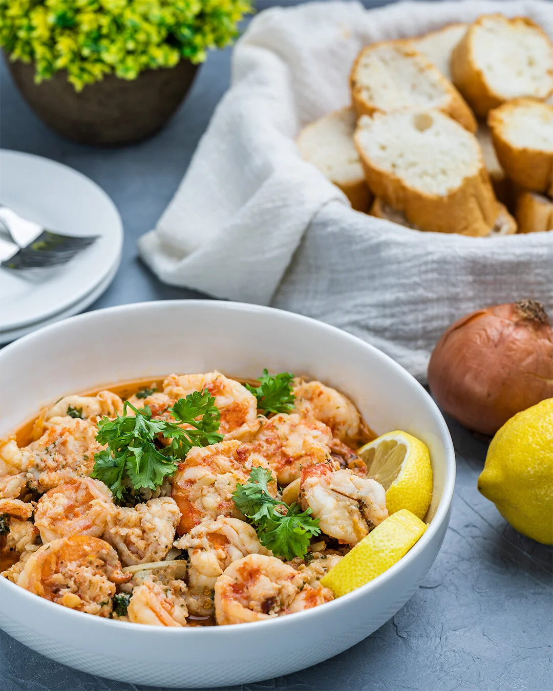 A bowl of shrimp in flavorful sauce garnished with fresh parsley and lemon wedges, with a basket of sliced bread and some lemon, onion, and greenery in the background.