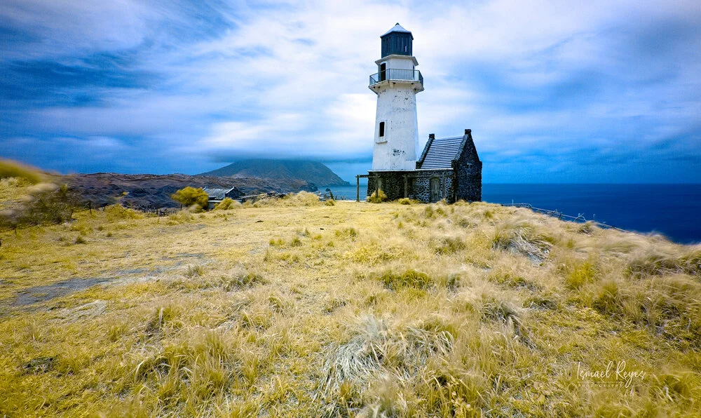 A lighthouse on a grassy cliff overlooking the ocean under a partly cloudy sky, with a mountain in the distance.