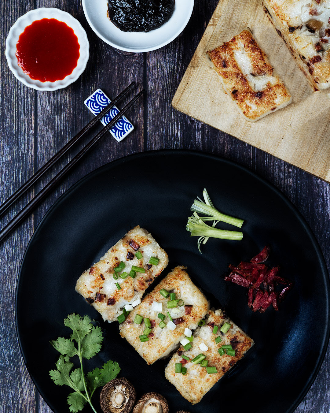 Pan-fried fish cakes garnished with chopped green onions on a black plate, accompanied by celery sticks, pickled beetroot, and mushrooms, with dipping sauces and other Chinese dishes on the side.