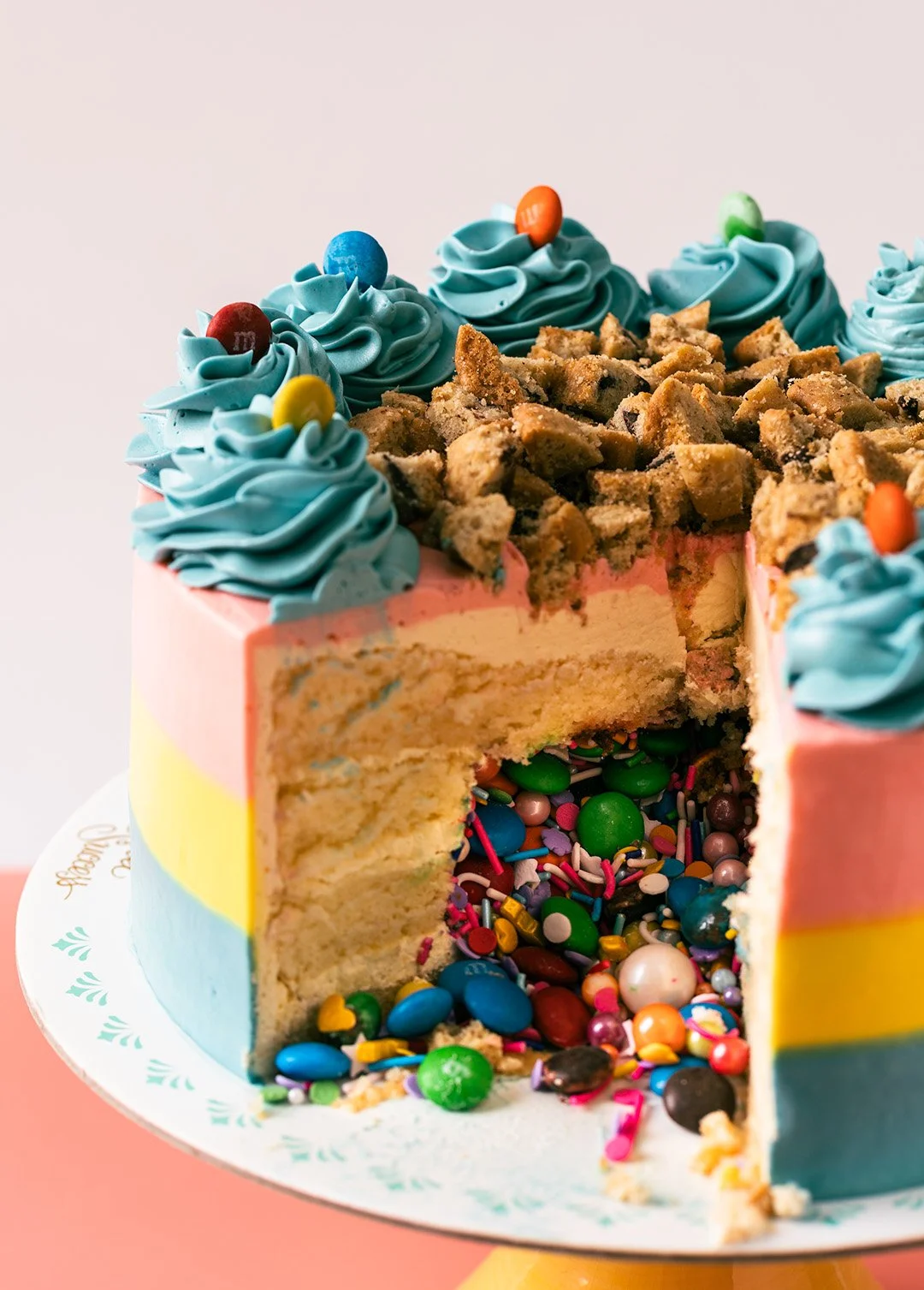 A colorful birthday cake with blue frosting, decorated with M&M candies, sprinkles, and cookie crumbles, with a slice cut out showing layers of cake and filling, on a pink and yellow surface.