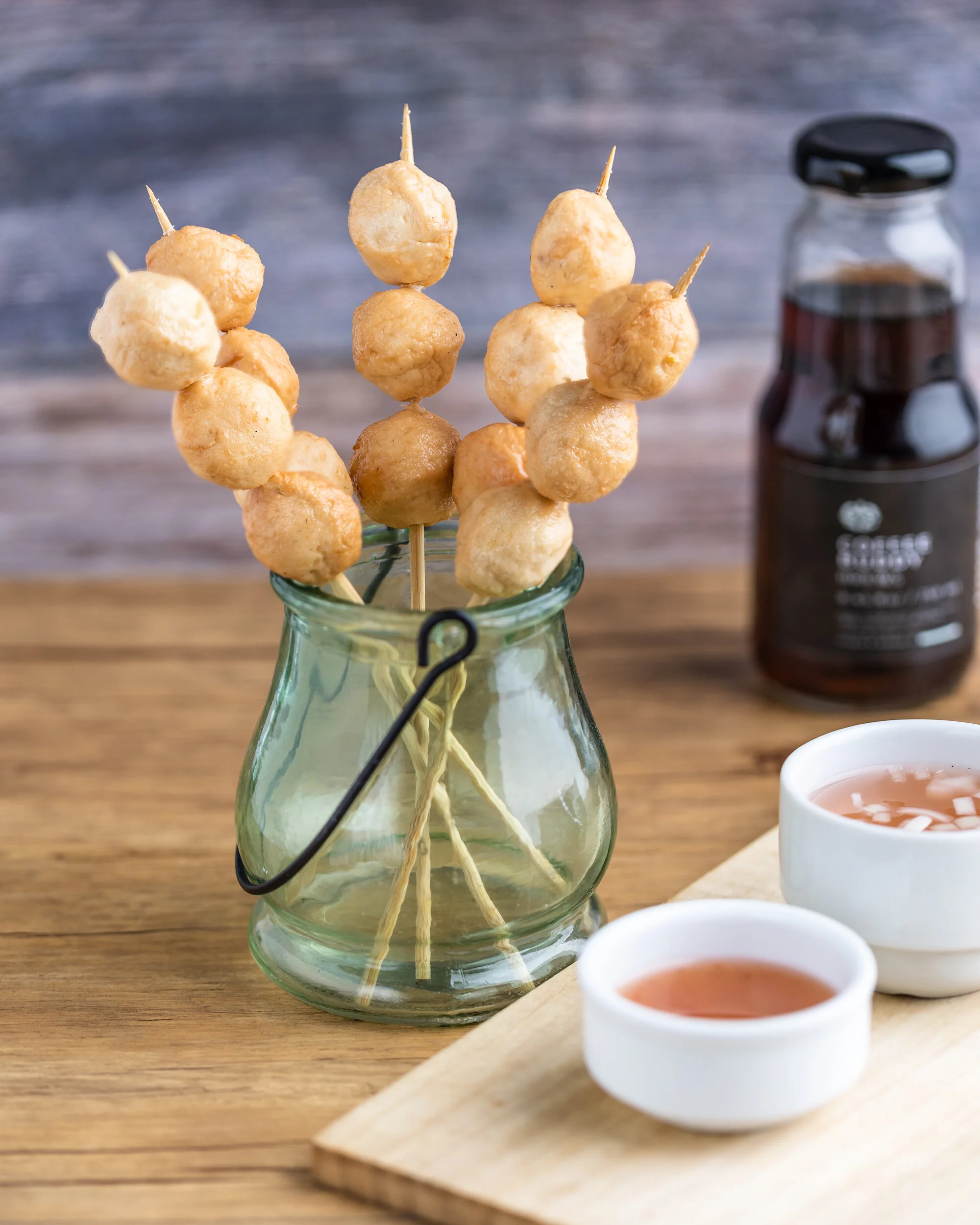 Skewers of fried meatballs in a glass jar, with small dishes of pink dipping sauce and a jar of sauce in the background, on a wooden surface.