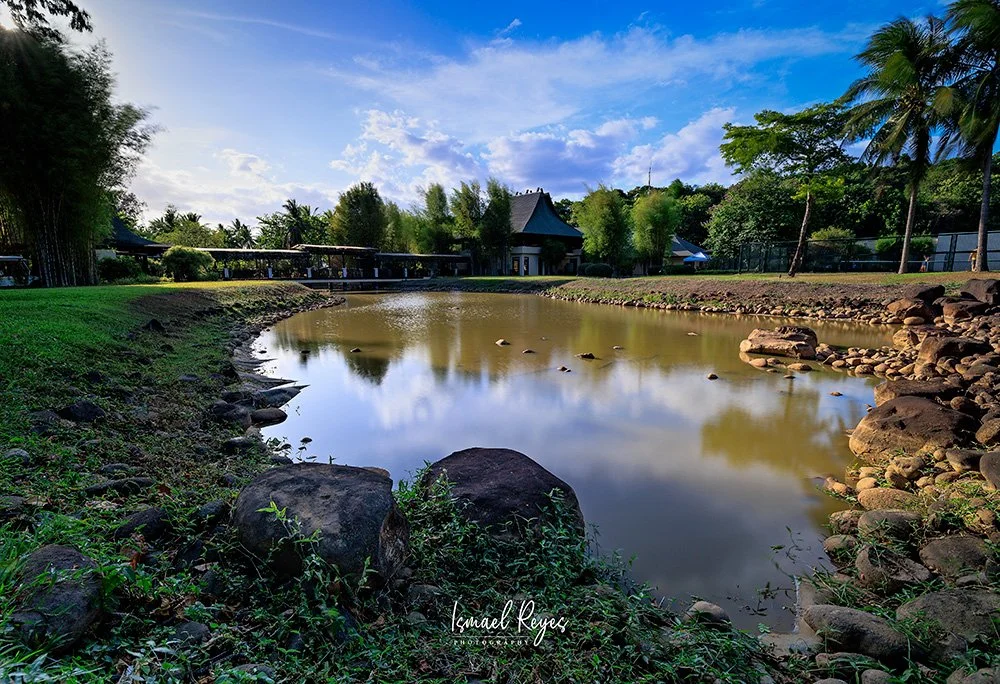 A tranquil pond surrounded by rocks and lush green trees with a partly cloudy sky overhead.