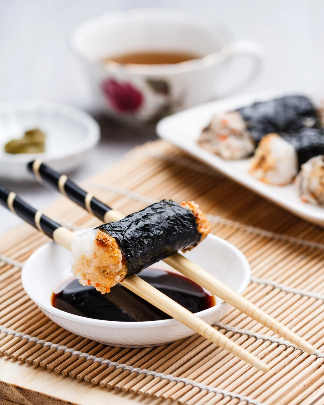 Close-up of a sushi roll with seaweed wrapping on chopsticks dipped in soy sauce. In the background, there is a plate with more sushi, a cup of tea, and small bowls containing wasabi and pickled ginger, all placed on a bamboo mat.