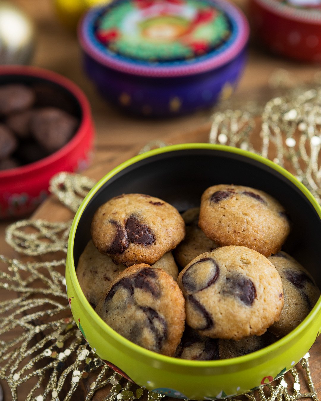 A tin of chocolate chip cookies with a festive background of colorful tins and gold decorations.