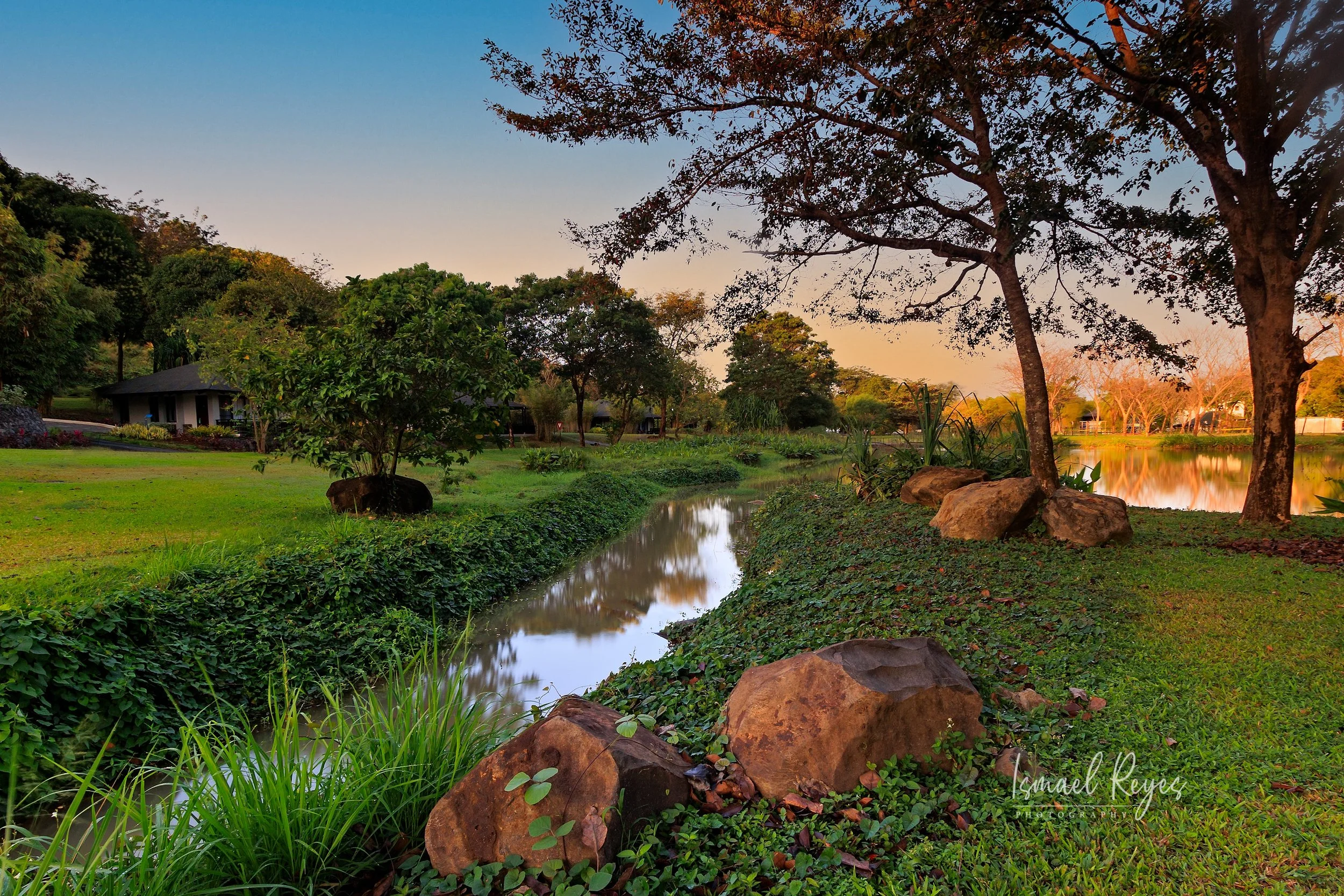 Scenic park with a small river, large rocks along the riverbank, green grass, trees, and a building in the background during sunset or sunrise.