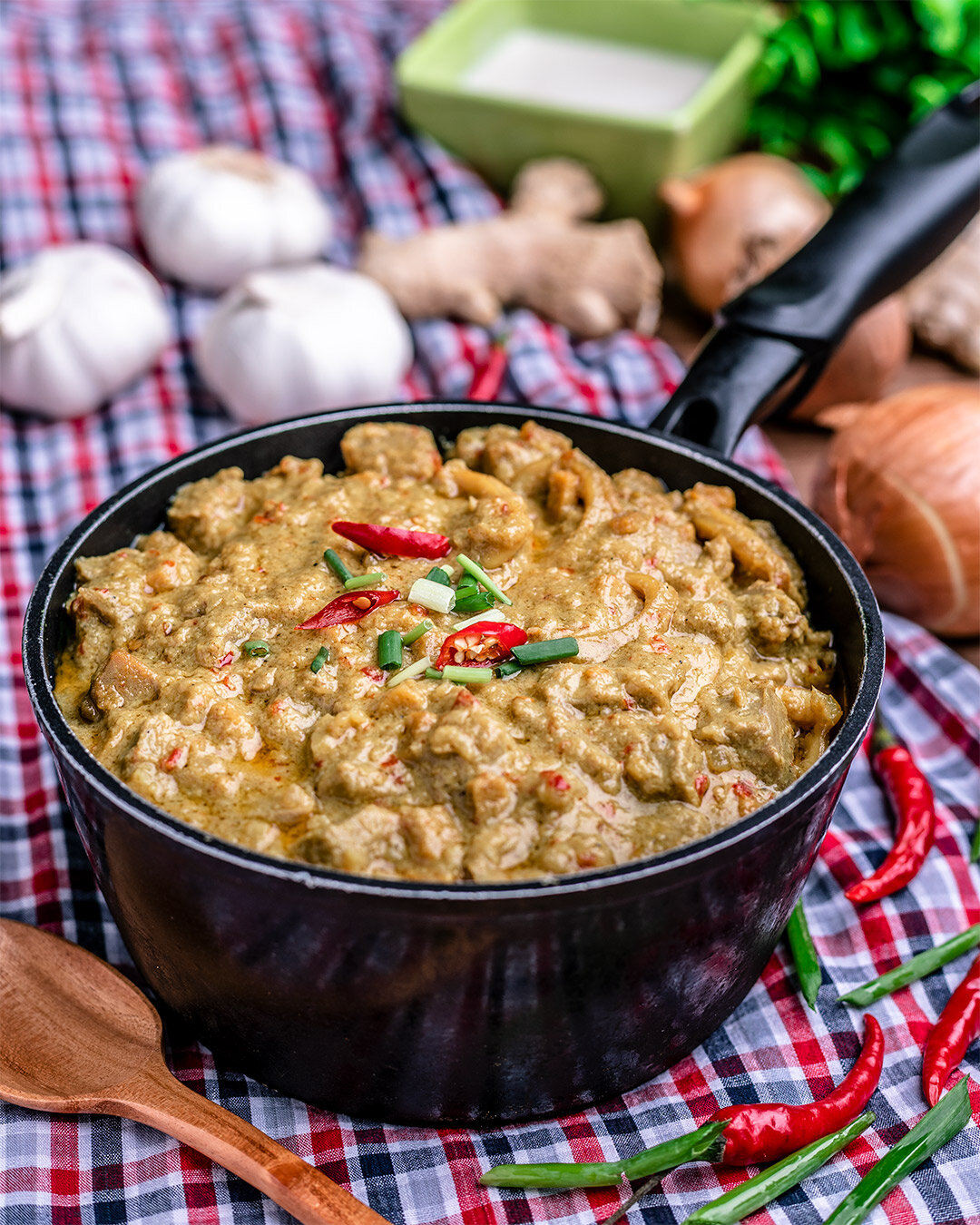 A black skillet filled with a creamy chicken curry garnished with sliced red chili peppers and chopped green onions, placed on a red and white checkered tablecloth with garlic, onions, ginger, and a green container in the background.