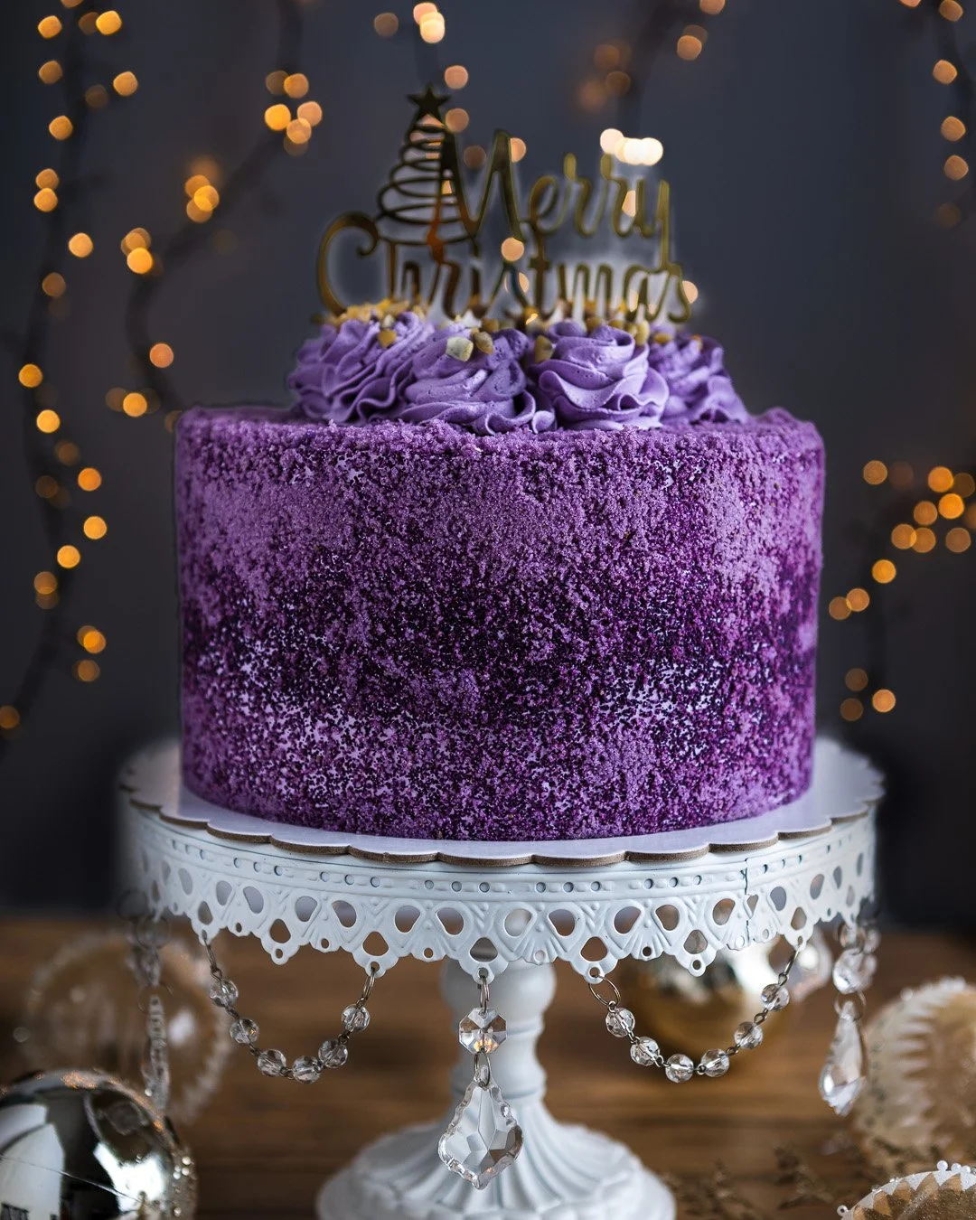 A Christmas cake with purple icing and sprinkles on a decorative white cake stand, topped with purple frosting rosettes and a gold 'Merry Christmas' topper, against a dark background with warm bokeh lights.