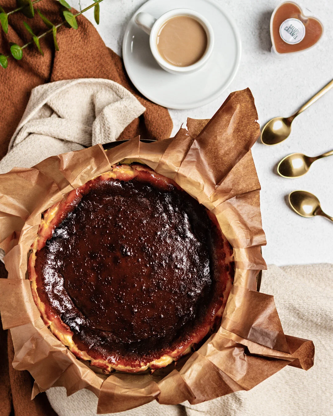 A burnt cheesecake on parchment paper, a cup of coffee with cream, and a small bottle of caramel sauce on a white surface, with three gold spoons and some folded cloths nearby.