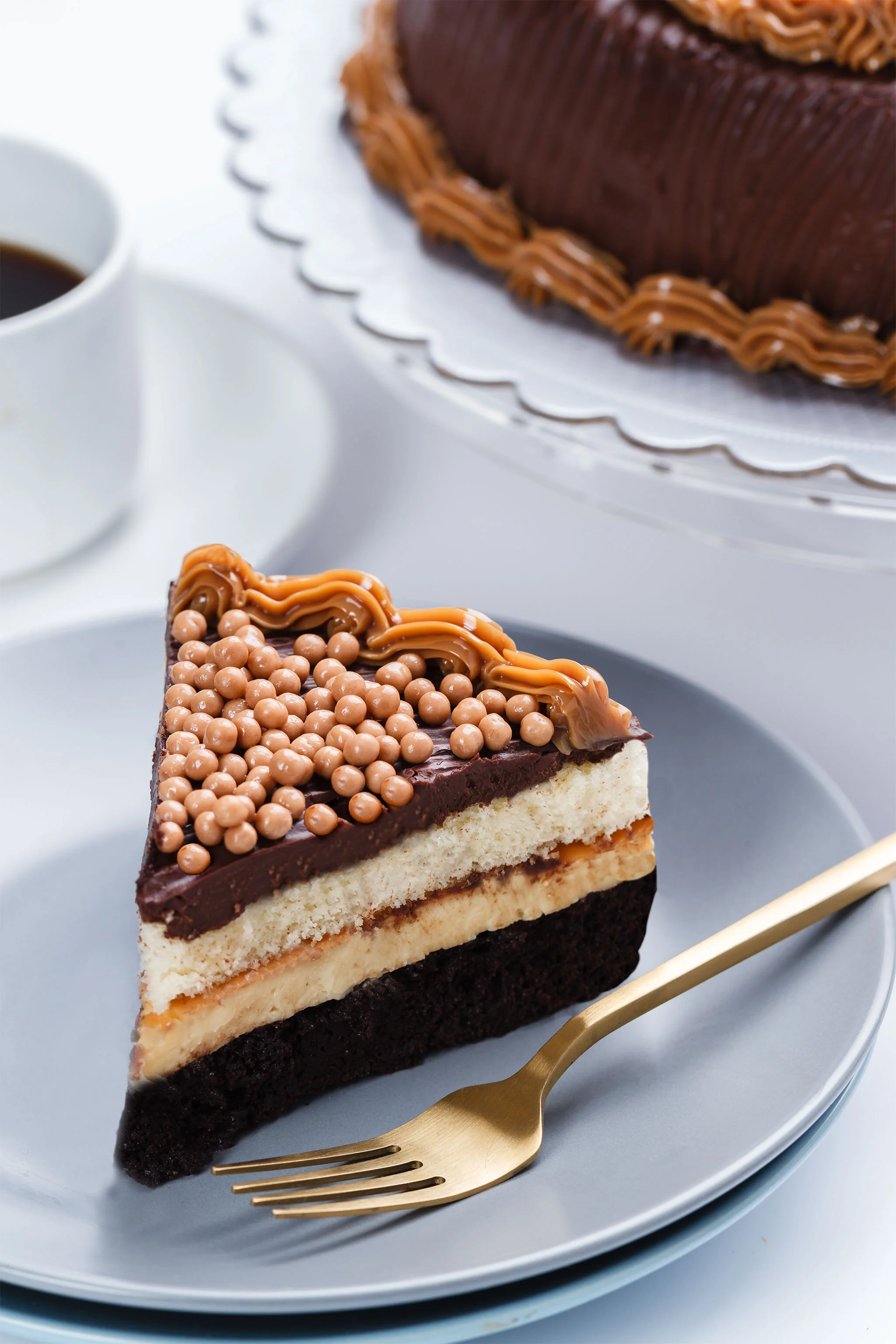 A slice of layered cake on a gray plate, topped with chocolate glaze, caramel drizzle, and small round caramel-colored candies, with a cup of coffee in the background.