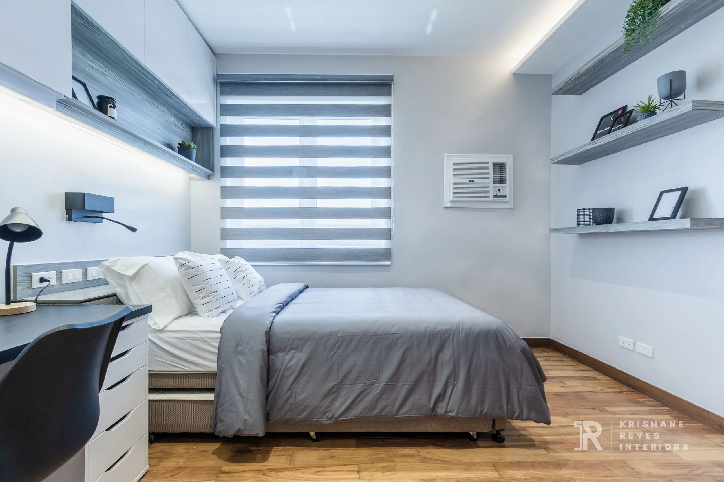 A modern, minimalist bedroom with a bed, desk, and shelves. The bed has white pillows and gray bedding, positioned near a window with horizontal blinds. The room features white walls, wooden flooring, and decorative accessories like framed photos and
