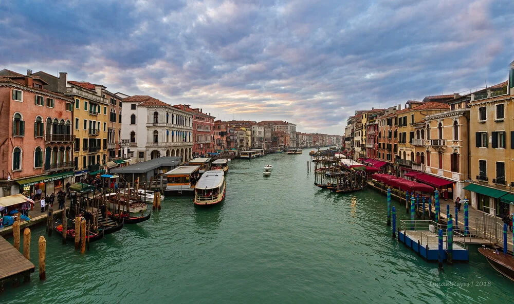 Colorful buildings line both sides of a canal in Venice, Italy, with boats and gondolas docked along the water and a cloudy sky overhead.
